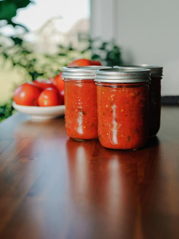 Three jars of homemade tomato sauce on a wooden table, with a bowl of fresh tomatoes and green leaves in the background—perfect inspiration if you’re looking for a homemade salsa recipe to enjoy all year round.