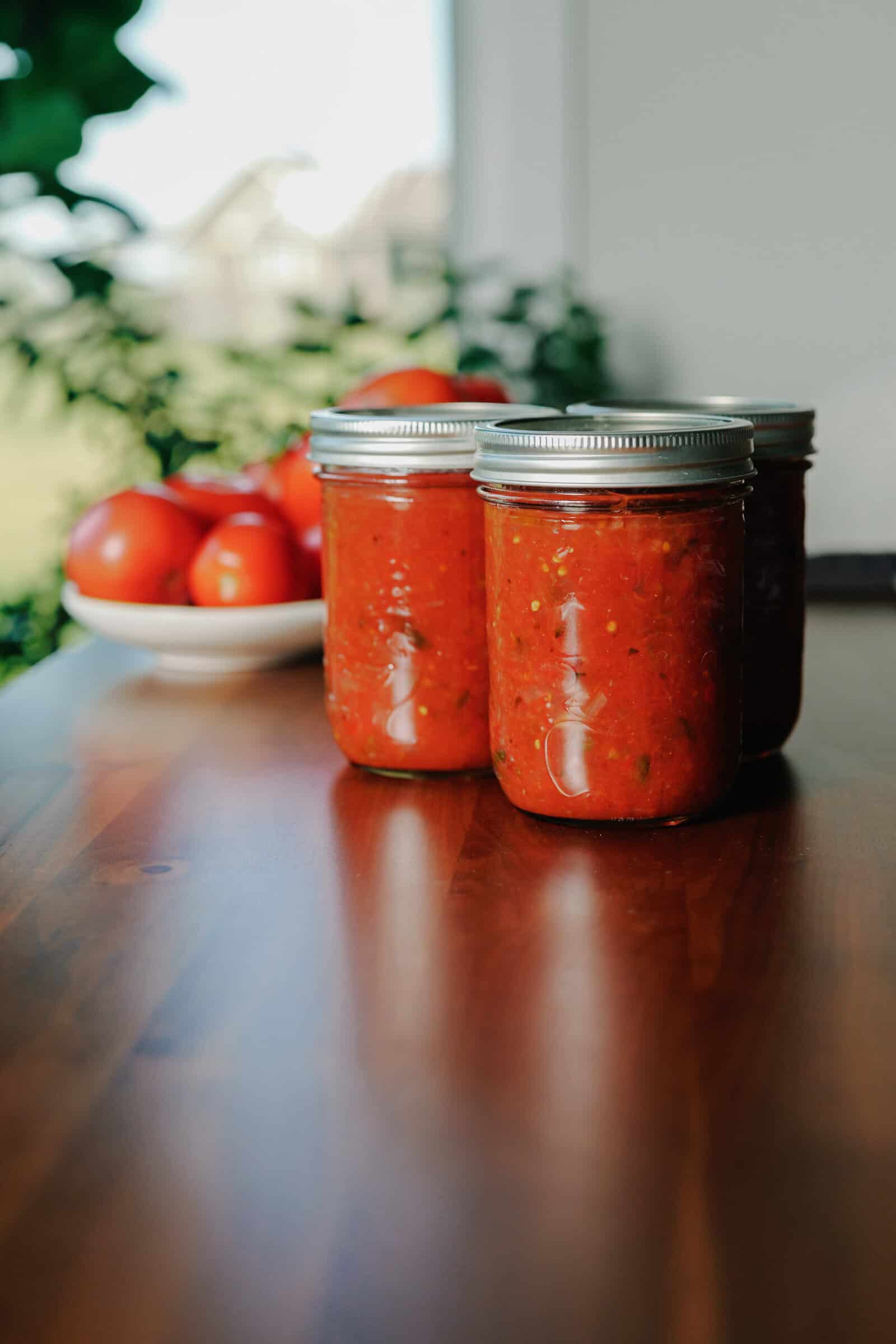 Three jars of homemade tomato sauce on a wooden table, with a bowl of fresh tomatoes and green leaves in the background—perfect inspiration if you’re looking for a homemade salsa recipe to enjoy all year round.