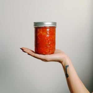A hand with a key tattoo on the wrist holds a glass jar filled with vibrant red homemade salsa, showcasing the results of a classic canned salsa recipe against a plain background.