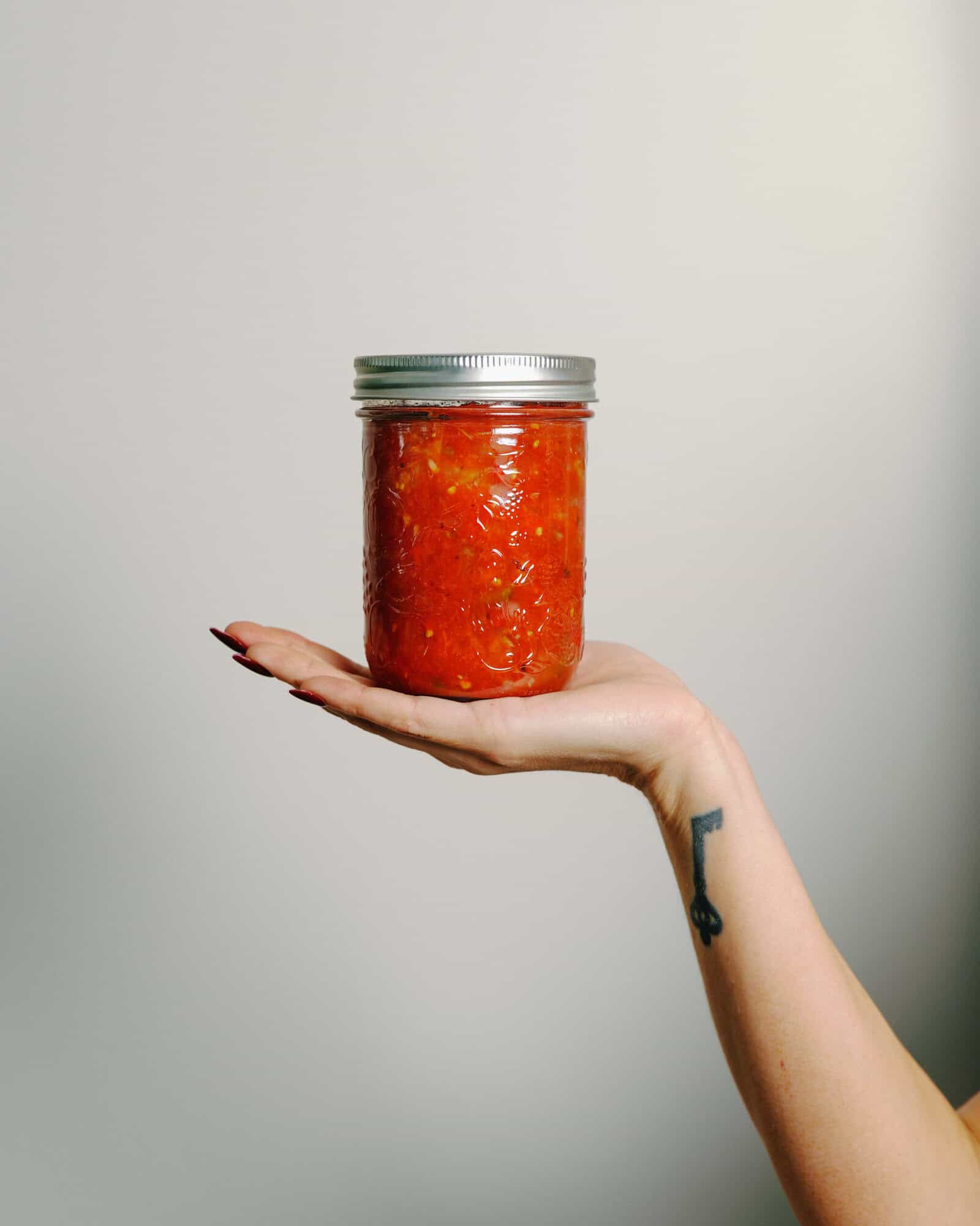 A hand with a key tattoo on the wrist holds a glass jar filled with vibrant red homemade salsa, showcasing the results of a classic canned salsa recipe against a plain background.