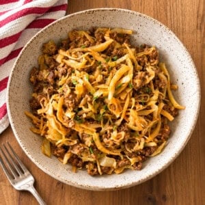 A white bowl filled with a savory cabbage hamburger helper dish, garnished with chopped herbs, sits on a wooden table next to a beige cloth napkin and a pan with more of the hearty hamburger helper recipe.