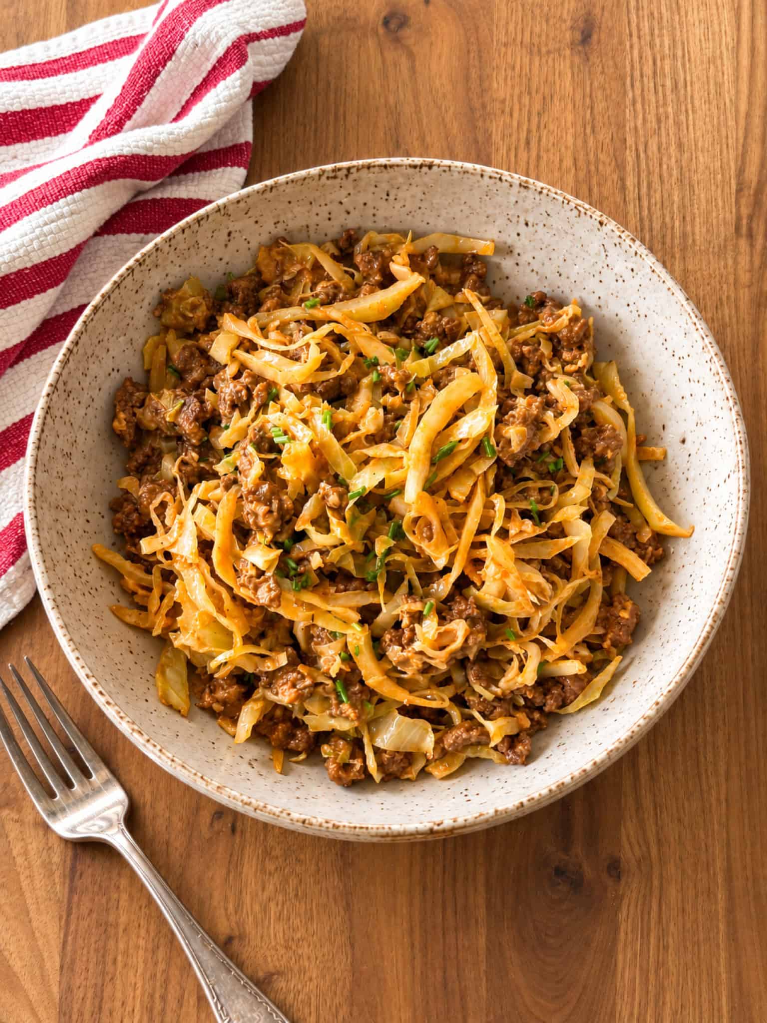 A white bowl filled with a savory cabbage hamburger helper dish, garnished with chopped herbs, sits on a wooden table next to a beige cloth napkin and a pan with more of the hearty hamburger helper recipe.