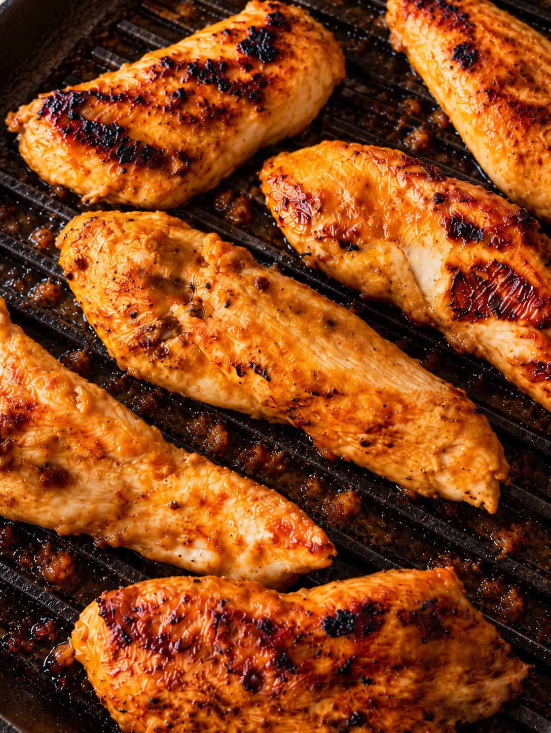 Several seasoned, golden-brown yogurt marinated chicken breast strips cooking in a ridged grill pan.