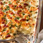 A glass baking dish containing cheesy scalloped potatoes in a creamy sauce with melted cheese and chives, with a portion scooped out by a serving spoon.