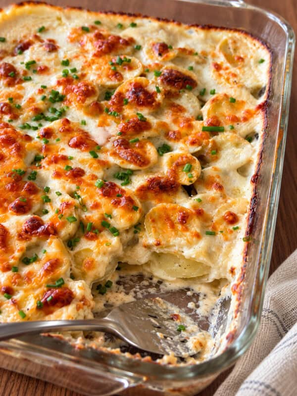 A glass baking dish containing cheesy scalloped potatoes in a creamy sauce with melted cheese and chives, with a portion scooped out by a serving spoon.