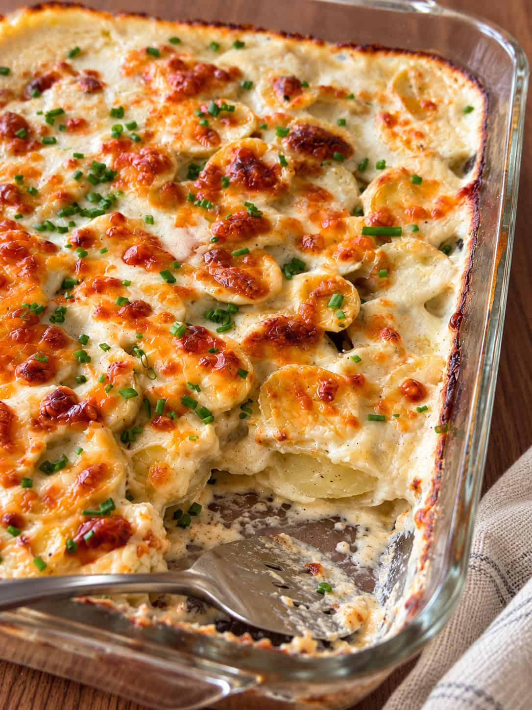 A glass baking dish containing cheesy scalloped potatoes in a creamy sauce with melted cheese and chives, with a portion scooped out by a serving spoon.