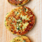 Three golden-brown cheese and chive stuffed potato cakes garnished with fresh herbs, arranged in a row on a wooden cutting board.