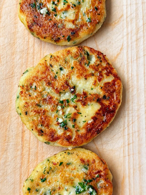 Three golden-brown cheese and chive stuffed potato cakes garnished with fresh herbs, arranged in a row on a wooden cutting board.