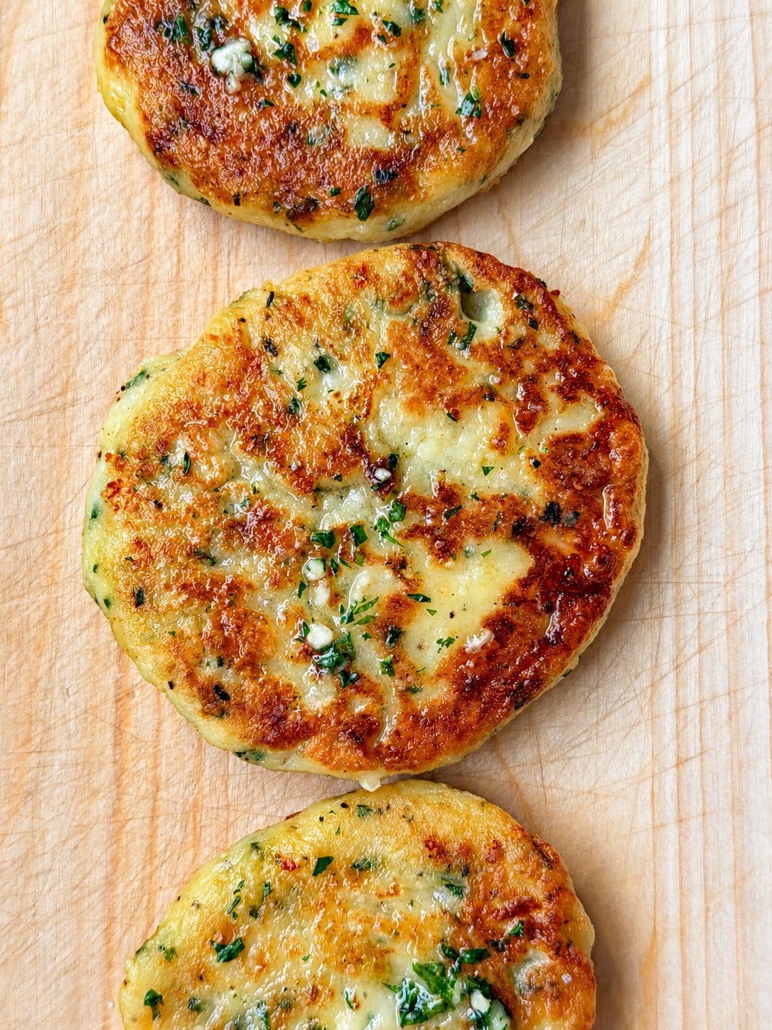 Three golden-brown cheese and chive stuffed potato cakes garnished with fresh herbs, arranged in a row on a wooden cutting board.