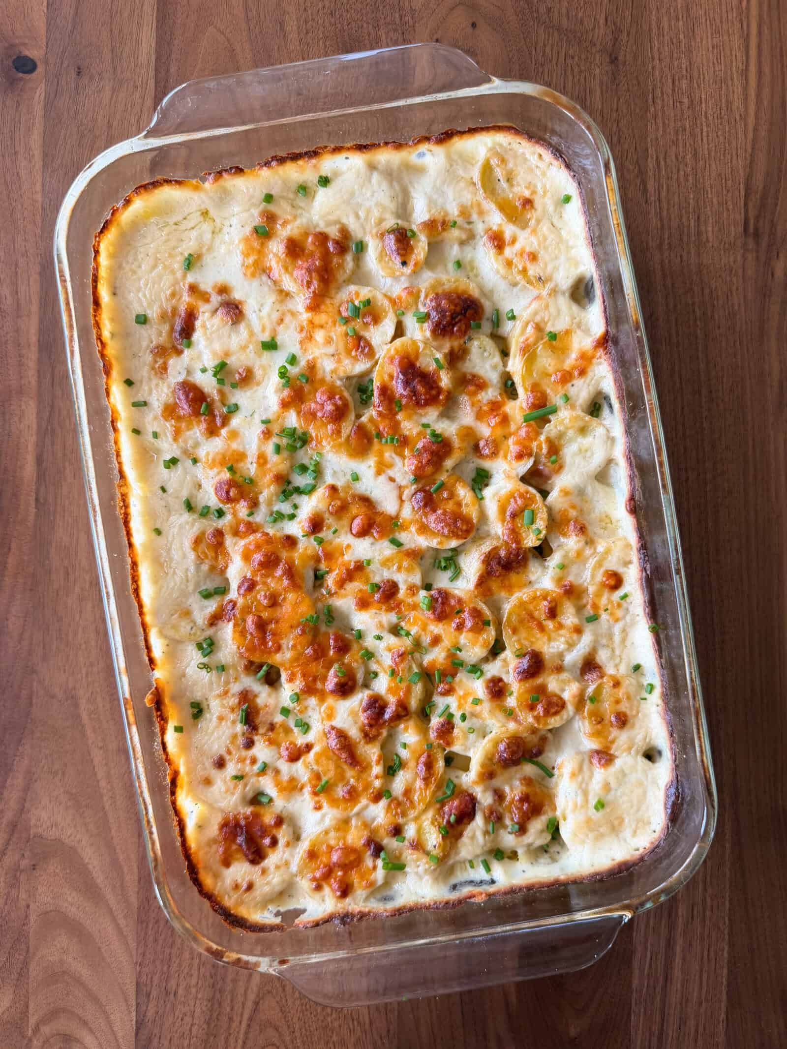 A glass baking dish filled with Boursin scalloped potatoes in a creamy sauce, topped with melted cheese and chopped chives.