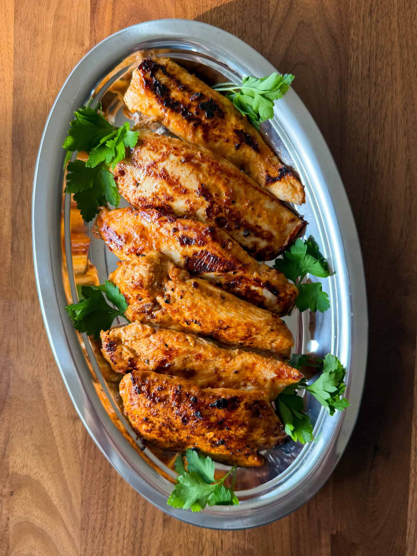 A silver platter filled with yogurt marinated chicken breast fillets, grilled and garnished with fresh parsley.