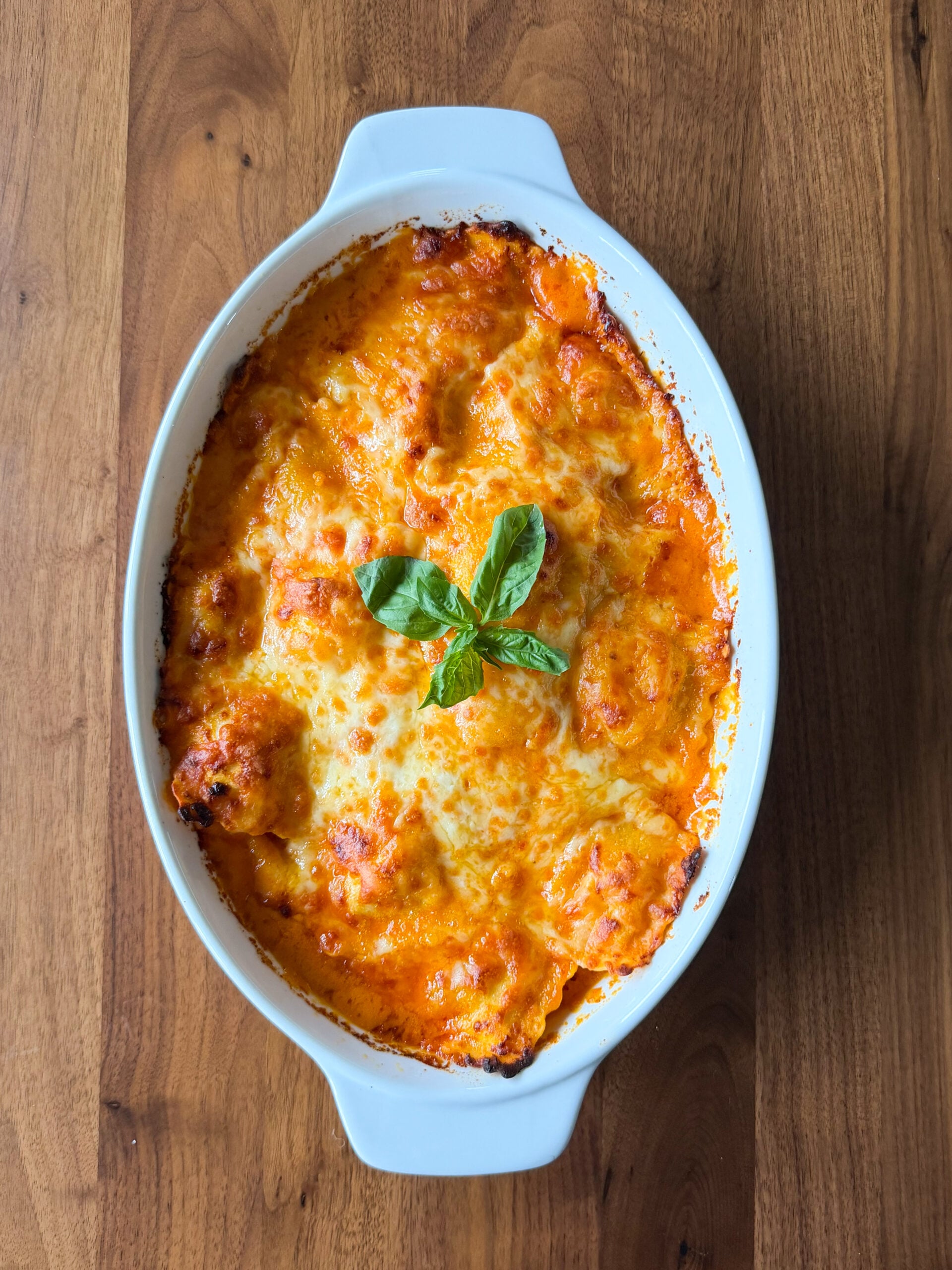 A baked ravioli dish with gochujang cream sauce, melted cheese, and fresh basil leaves in a white oval baking dish on a wooden surface.