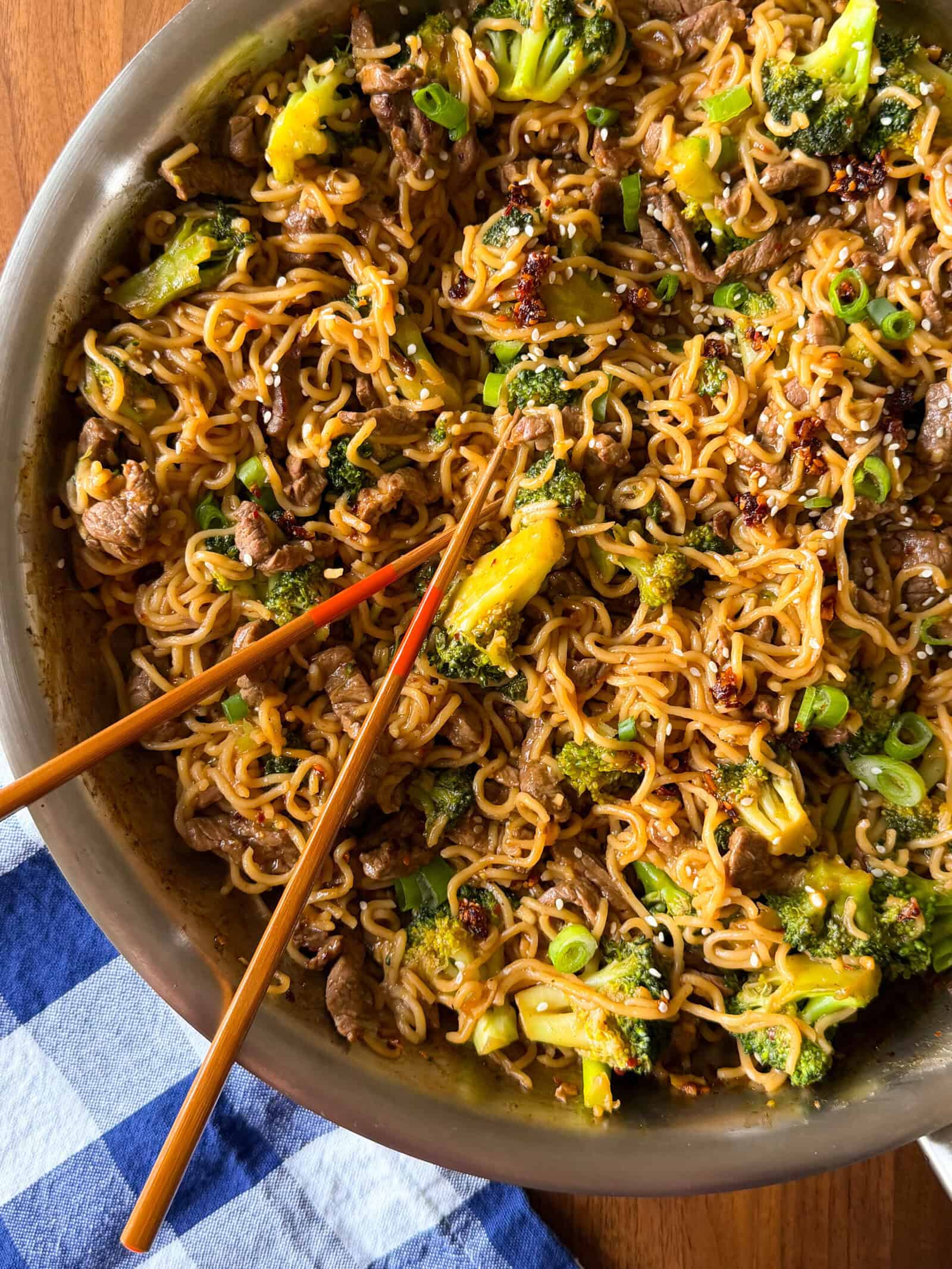 A skillet of beef and broccoli ramen with stir-fried noodles, broccoli, and green onions, with a pair of wooden chopsticks resting on top.