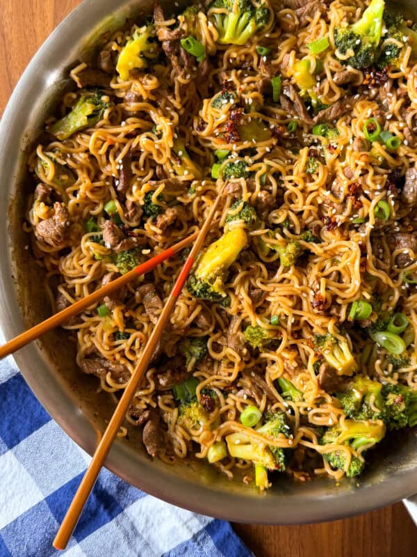 Beef and broccoli stir fry with noodles in a pan, topped with sesame seeds and green onions, with a pair of chopsticks resting on top.