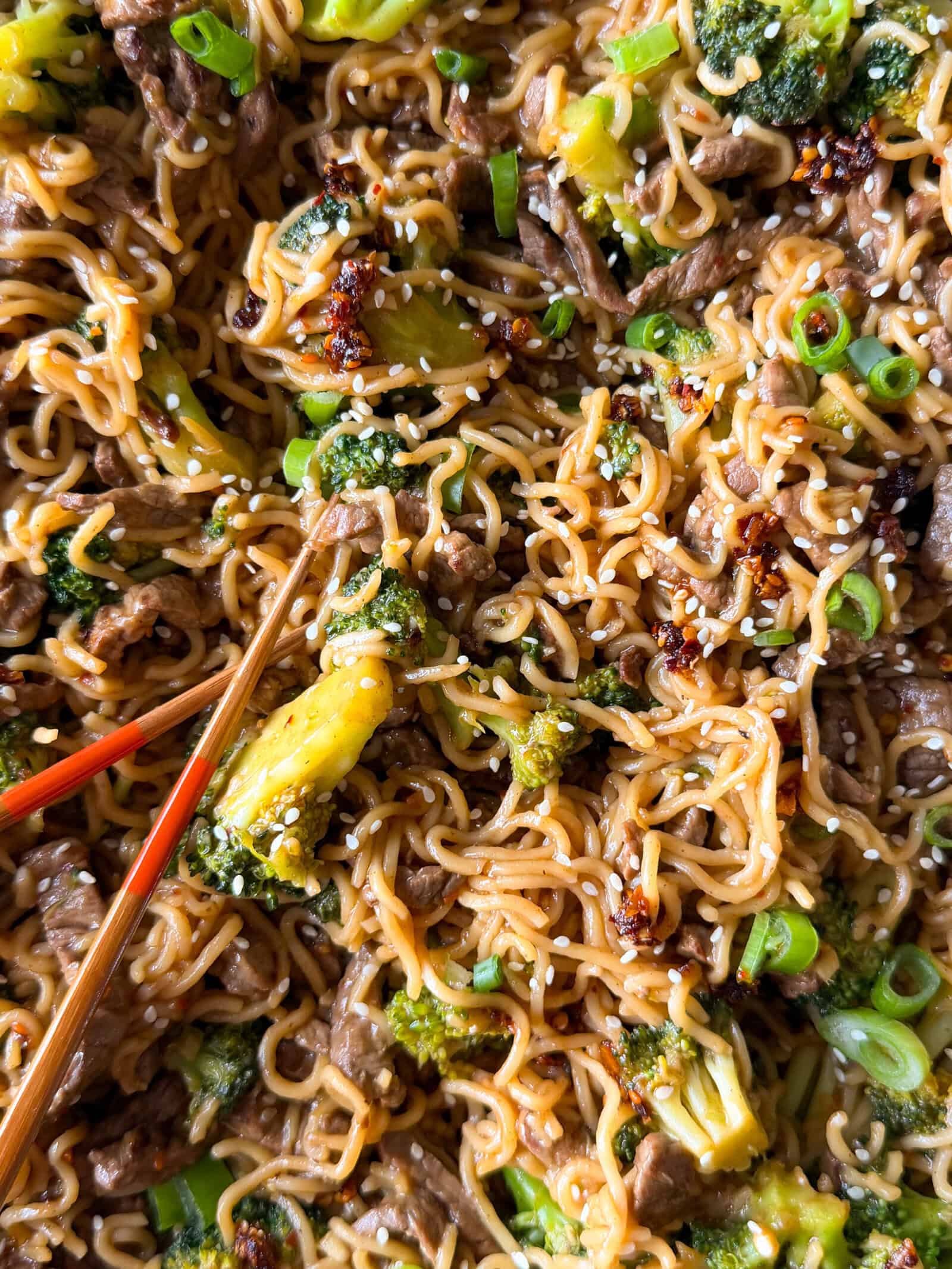 Beef and broccoli ramen stir-fried with broccoli and green onions, topped with sesame seeds and chili oil, with a pair of chopsticks resting in the dish.