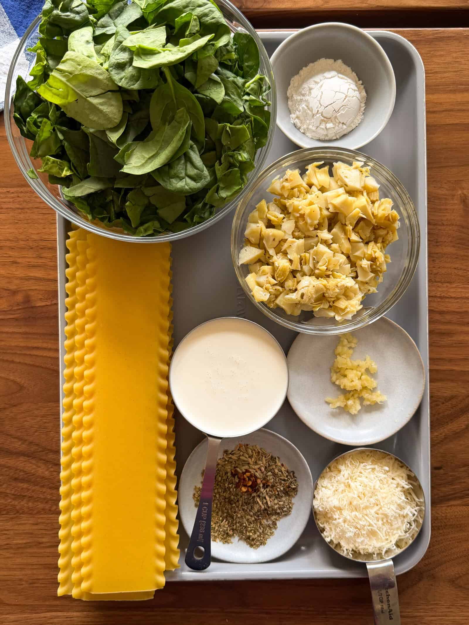 A tray of ingredients for White Lasagna Soup including a bowl of fresh spinach, lasagna noodles, chopped artichokes, a cup of cream, flour, minced garlic, spices, and shredded cheese.