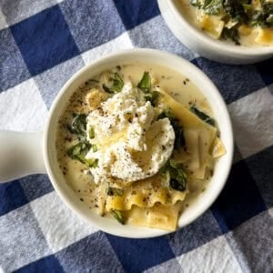 Two bowls of easy lasagna soup topped with dollops of ricotta, herbs, and pepper, resting on a blue and white checkered tablecloth.