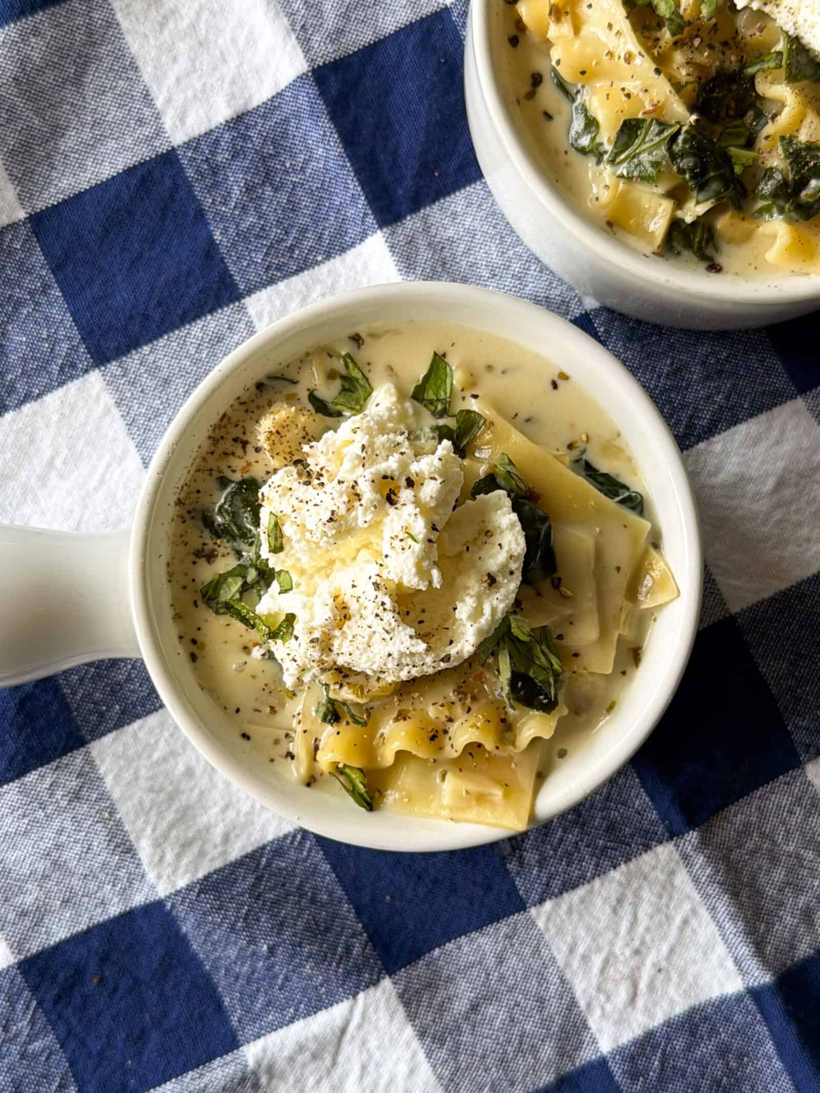 Two bowls of easy lasagna soup topped with dollops of ricotta, herbs, and pepper, resting on a blue and white checkered tablecloth.
