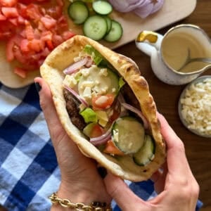 Hands holding a Middle Eastern inspired smashed pita sandwich filled with a burger patty, sliced cucumbers, tomatoes, red onions, and feta cheese, surrounded by bowls of the same fresh ingredients.