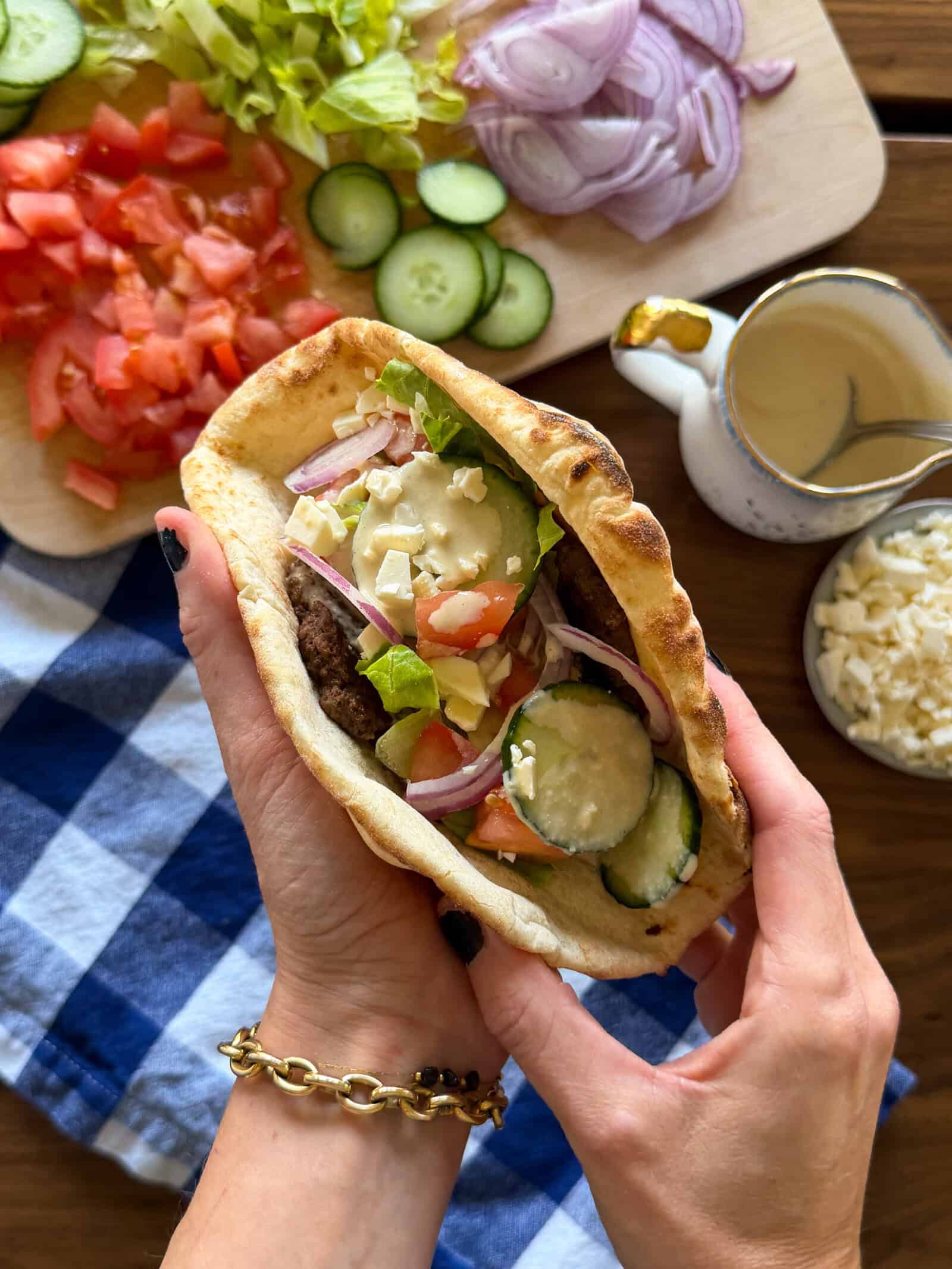 Hands holding a Middle Eastern inspired smashed pita sandwich filled with a burger patty, sliced cucumbers, tomatoes, red onions, and feta cheese, surrounded by bowls of the same fresh ingredients.