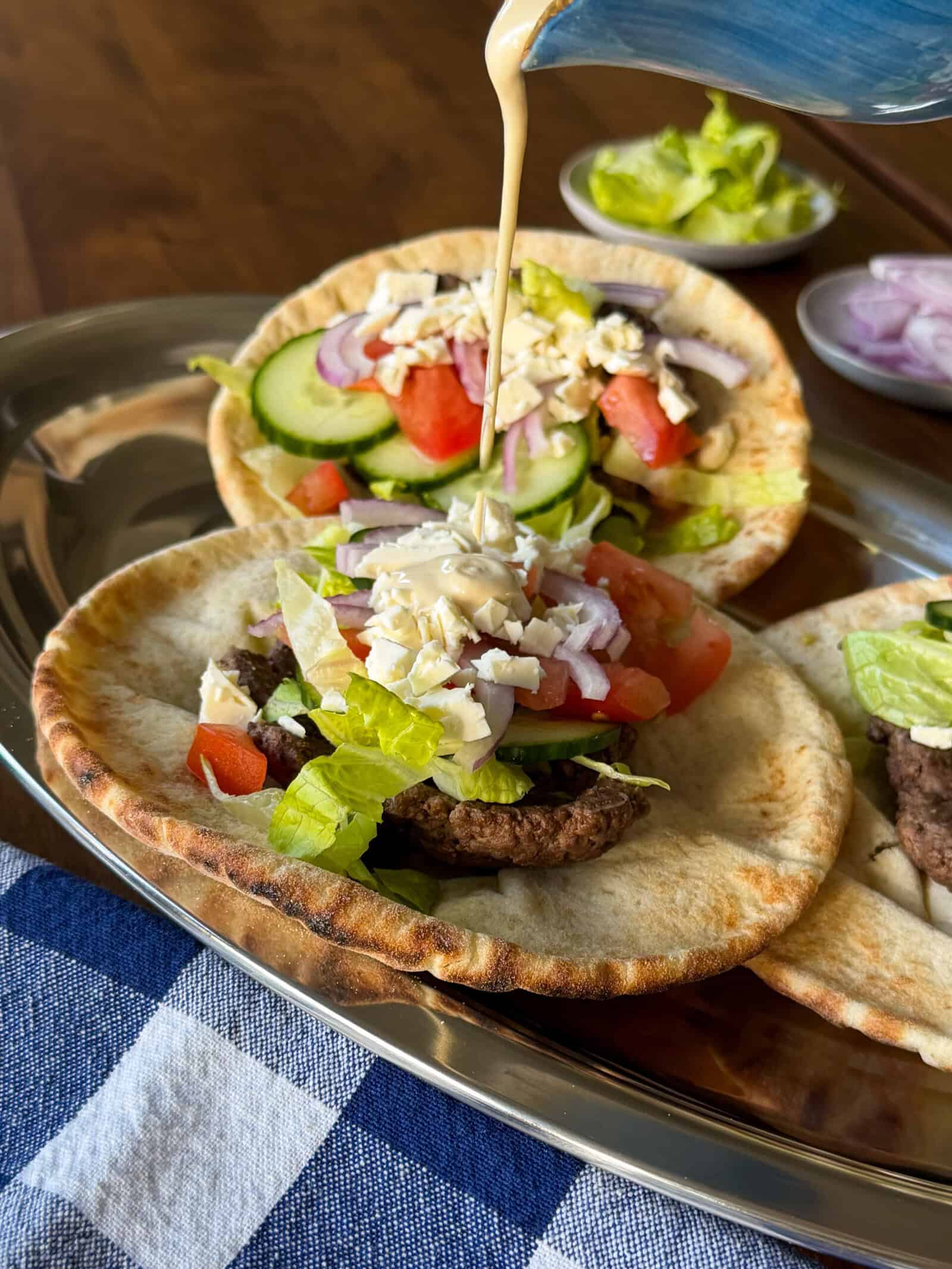 Creamy dressing being poured over a middle eastern inspired smashed pita taco topped with a burger patty, chopped vegetables, and crumbled cheese.