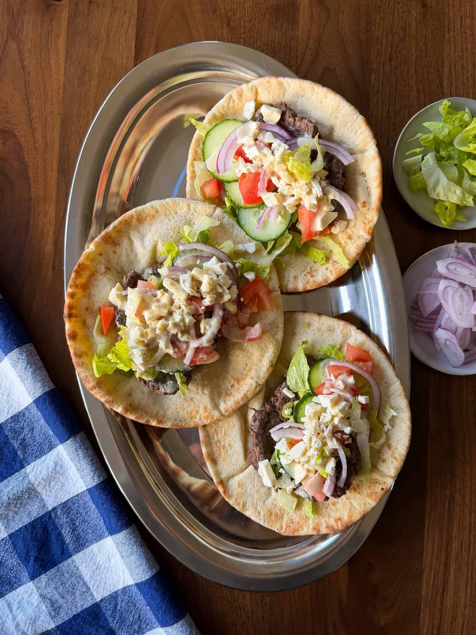 Three Middle Eastern inspired smashed pita sandwiches topped with meat, lettuce, cucumber, tomato, red onion, and crumbled cheese on an oval silver platter, with small side bowls of lettuce and onion.