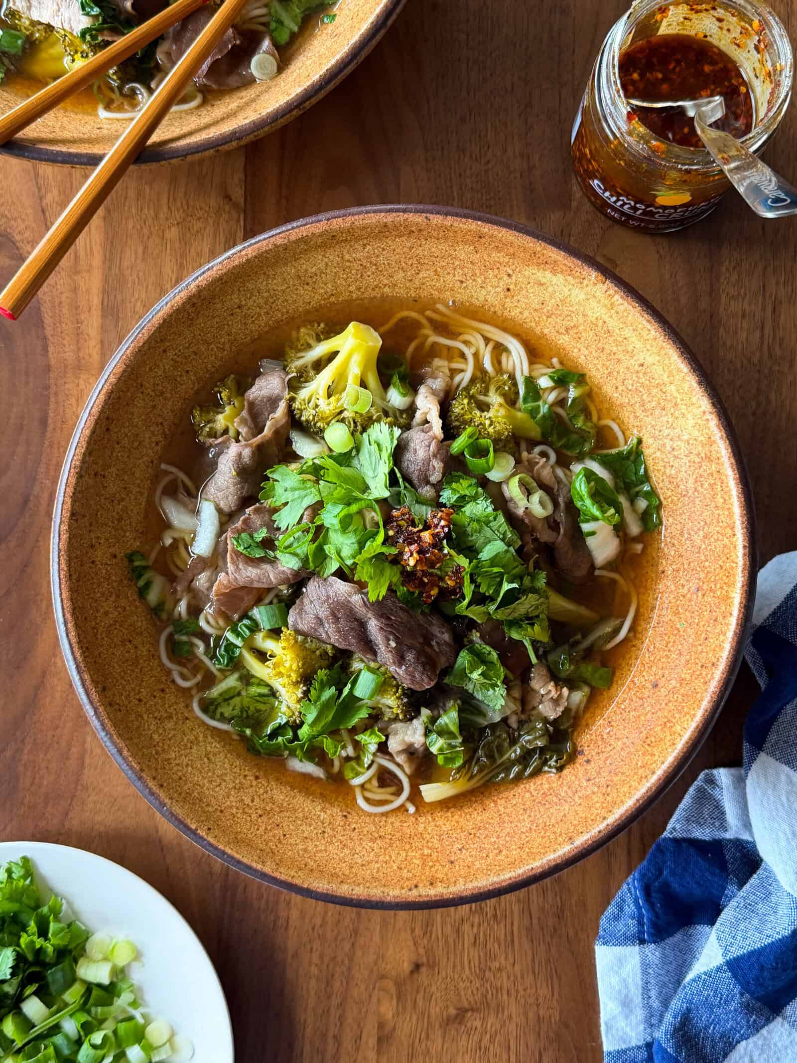 A bowl of Asian inspired beef and noodle soup topped with fresh herbs and chili oil, with a jar of chili crisp and side plates nearby.