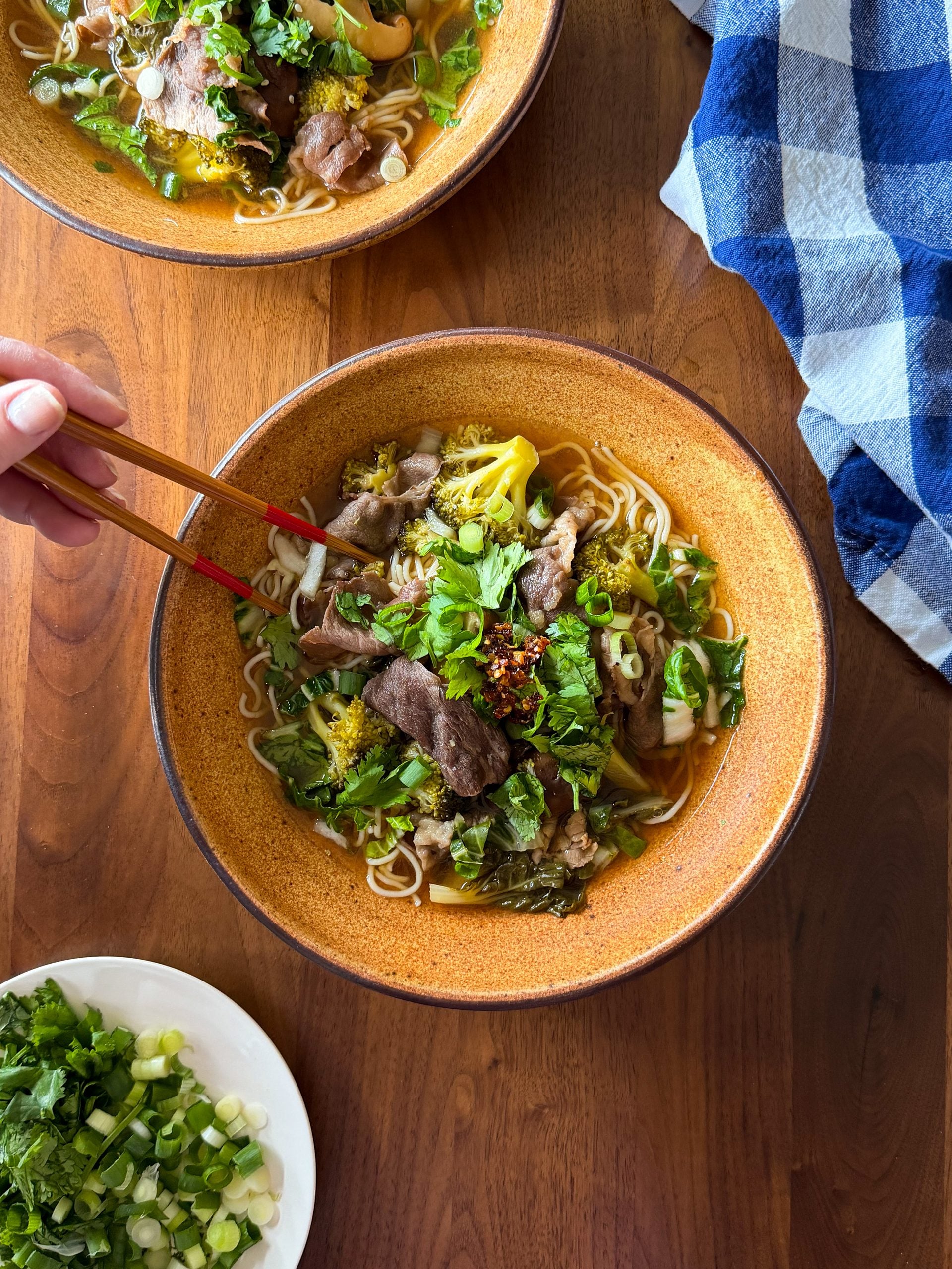 A person uses chopsticks to pick noodles from a bowl of Asian inspired beef and noodle soup, with a side plate of chopped cilantro and scallions on a wooden table.