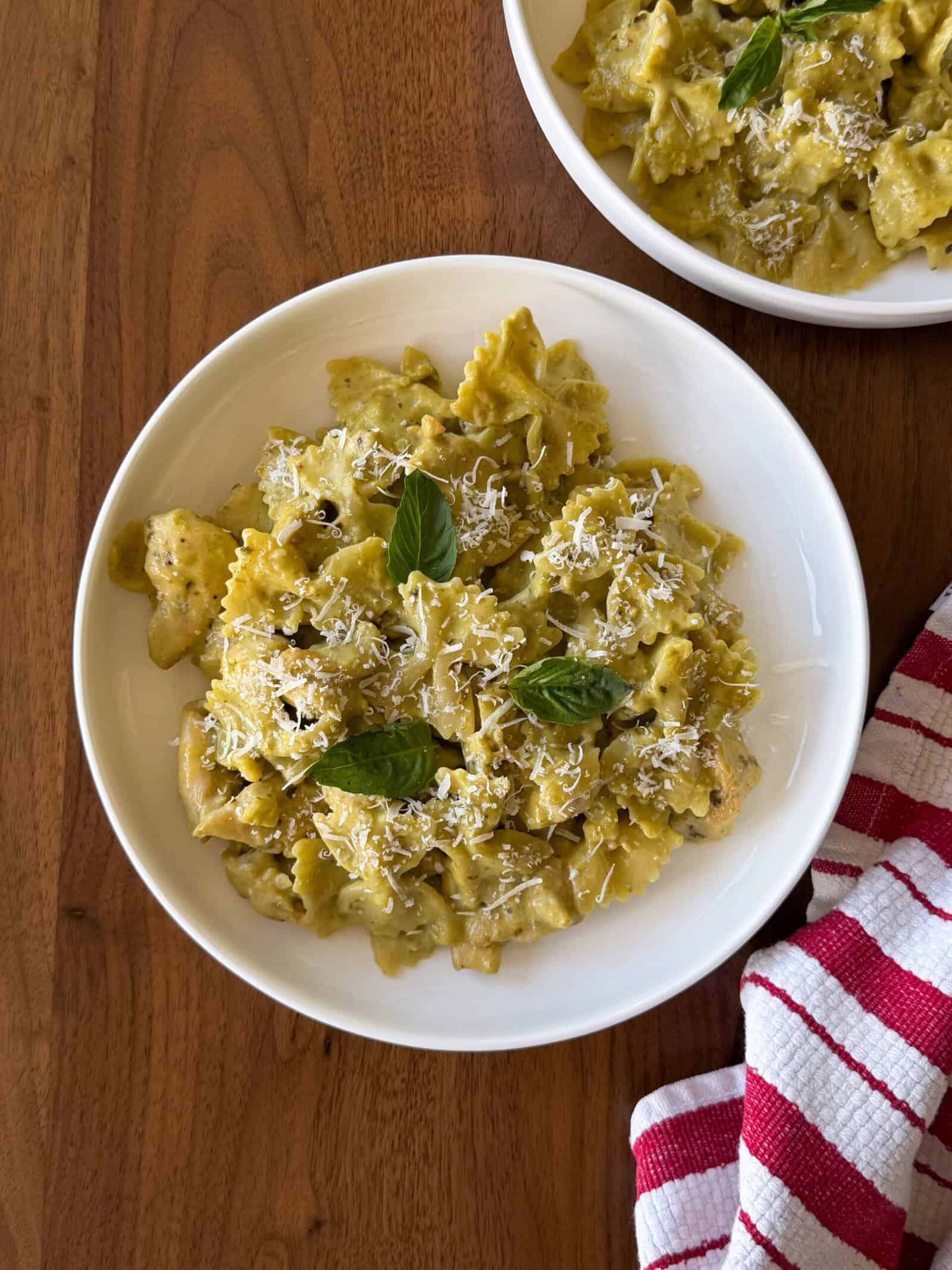 Bowtie chicken pesto pasta in a creamy green sauce, topped with grated cheese and fresh basil leaves, served in a white bowl.