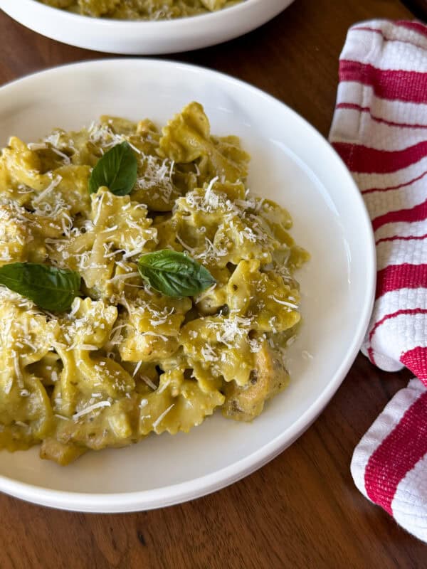 Chicken pesto pasta in a white bowl with green sauce, topped with grated cheese and fresh basil leaves next to a red and white striped kitchen towel.