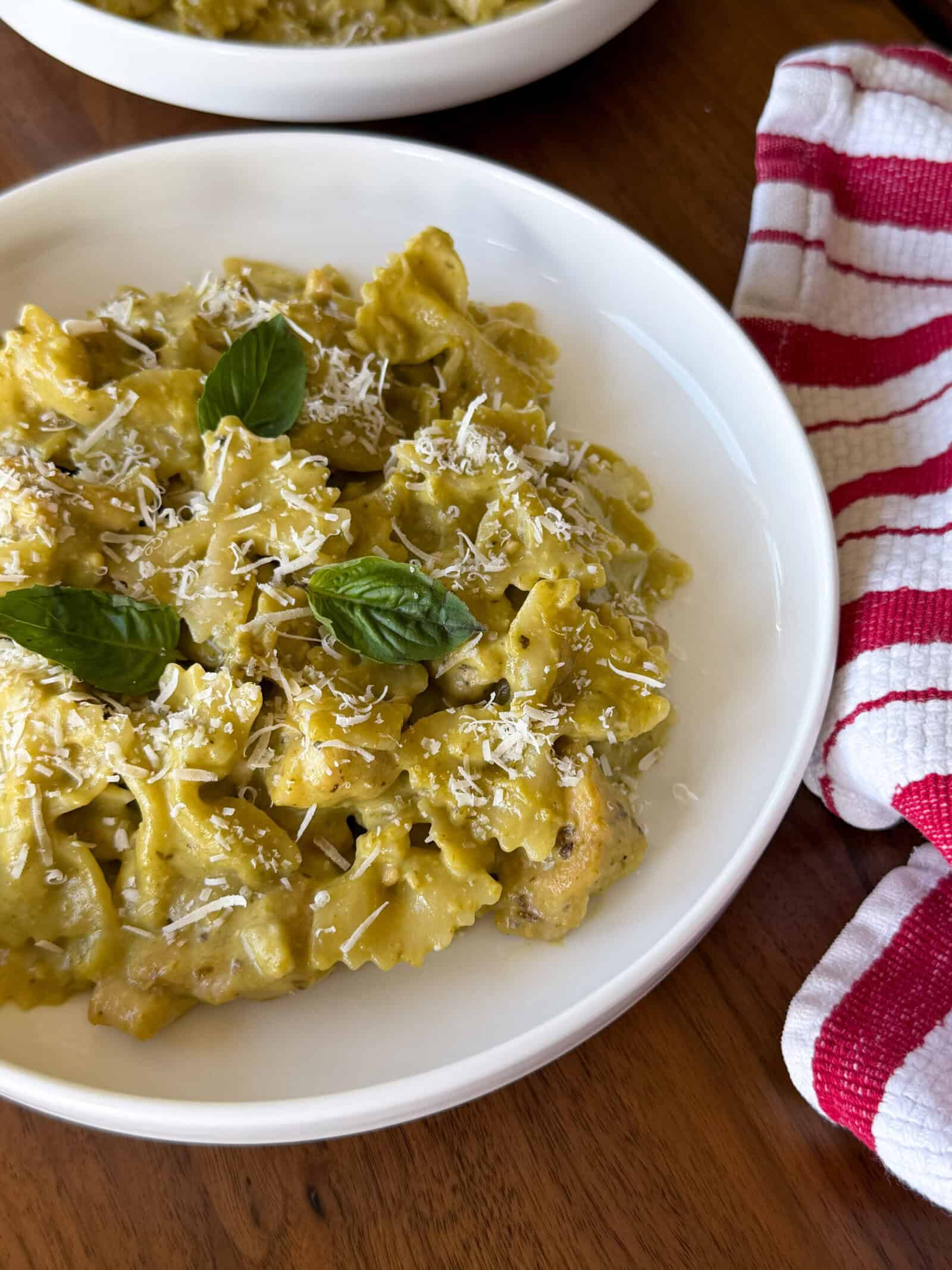 Chicken pesto pasta in a white bowl with green sauce, topped with grated cheese and fresh basil leaves next to a red and white striped kitchen towel.