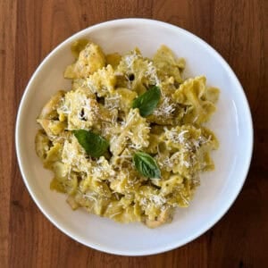 A bowl of farfalle chicken pesto pasta in creamy green sauce, topped with shredded cheese and fresh basil leaves.