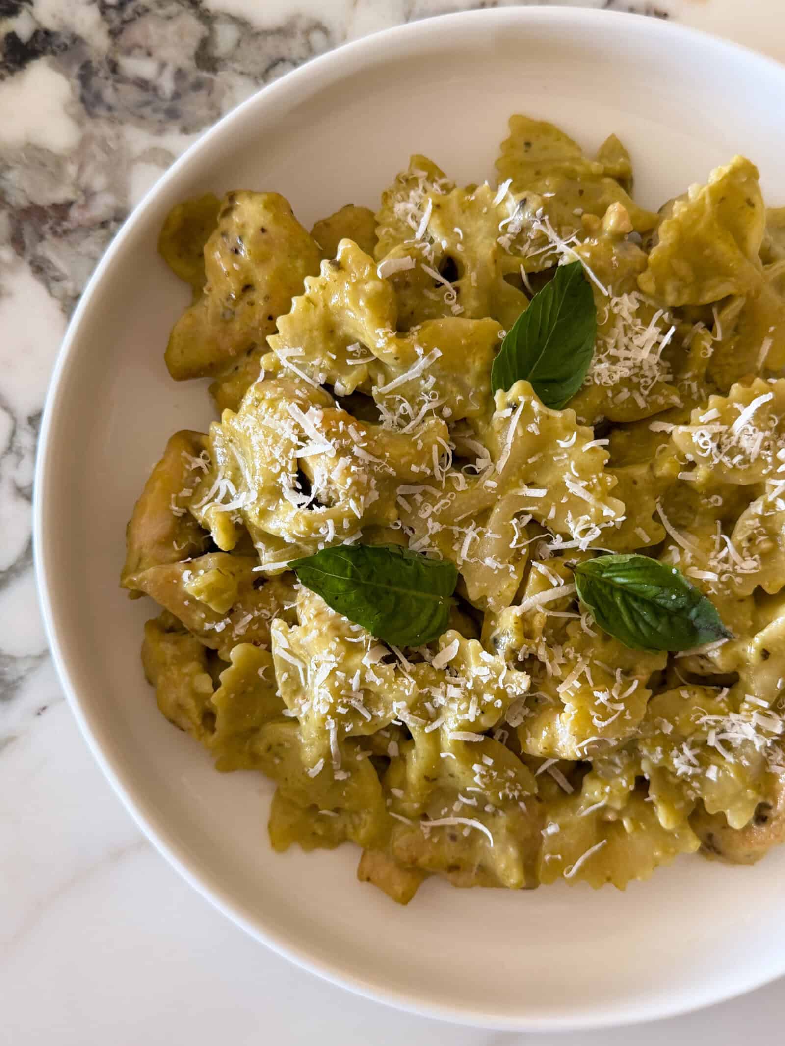 Farfalle chicken pesto pasta tossed in a creamy green sauce, topped with shredded cheese and fresh basil leaves in a white bowl.
