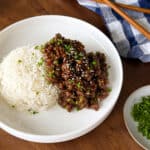 A plate of Sticky Honey Ginger Ground Beef over white rice topped with sesame seeds and chopped chives, served with a pair of chopsticks and a side of chives.