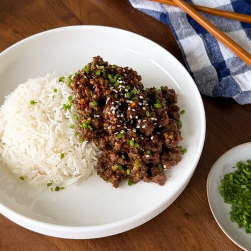 A plate of Sticky Honey Ginger Ground Beef over white rice topped with sesame seeds and chopped chives, served with a pair of chopsticks and a side of chives.