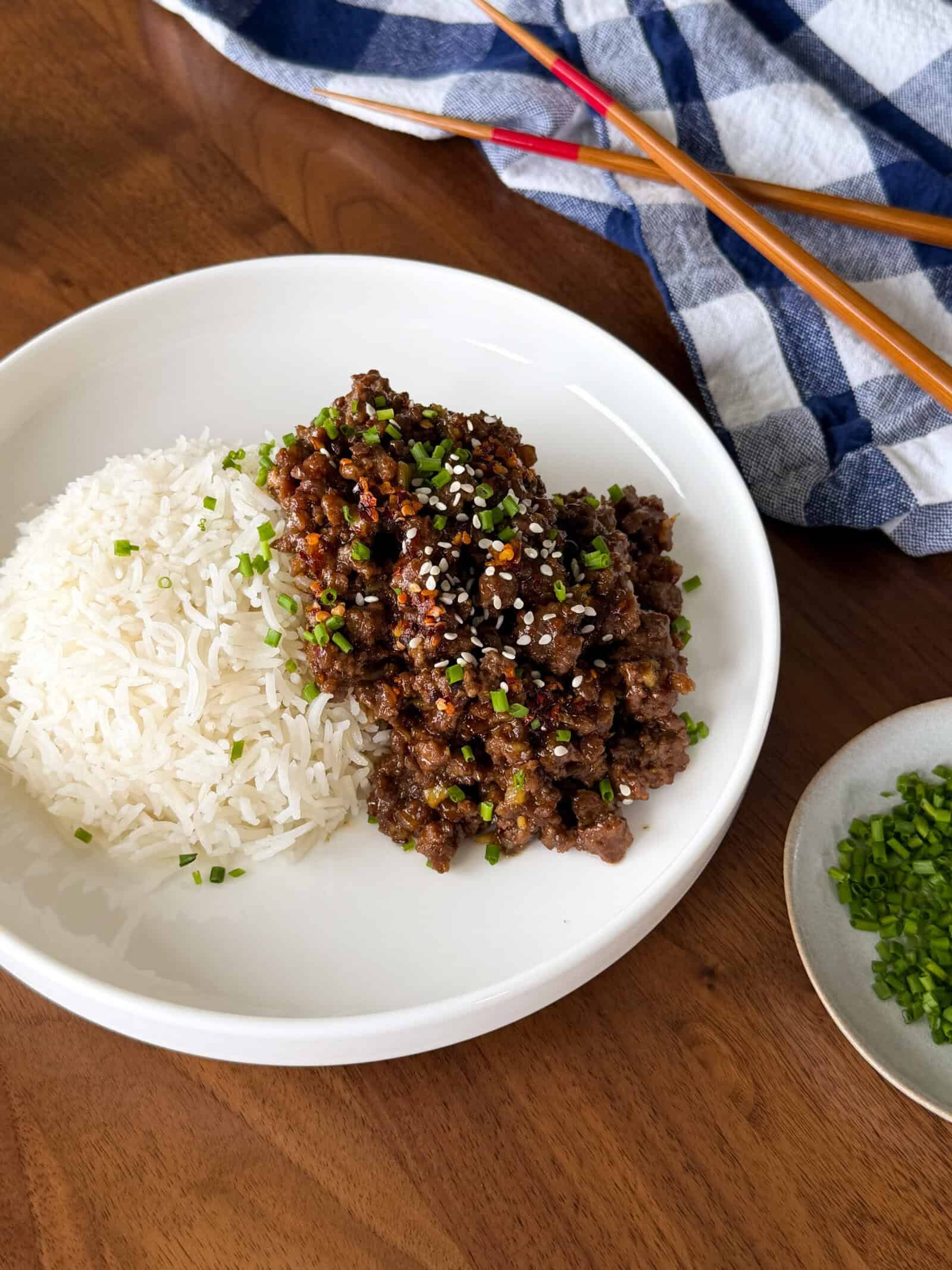 A plate of Sticky Honey Ginger Ground Beef over white rice topped with sesame seeds and chopped chives, served with a pair of chopsticks and a side of chives.