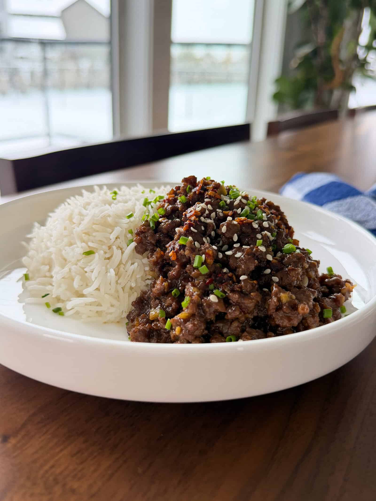 A bowl of sticky honey ginger ground beef topped with sesame seeds and chopped chives, served alongside a portion of white rice.