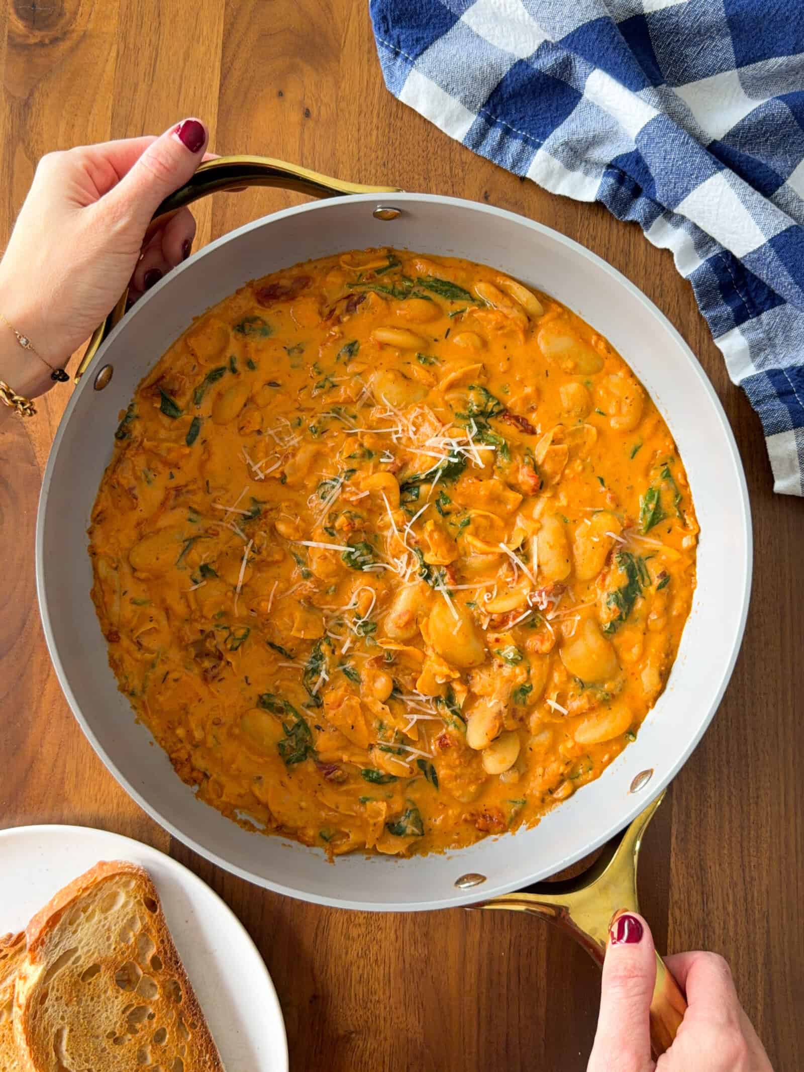 A pan of creamy Marry Me butter beans and spinach, held by two hands with red nail polish, next to a slice of toasted bread.