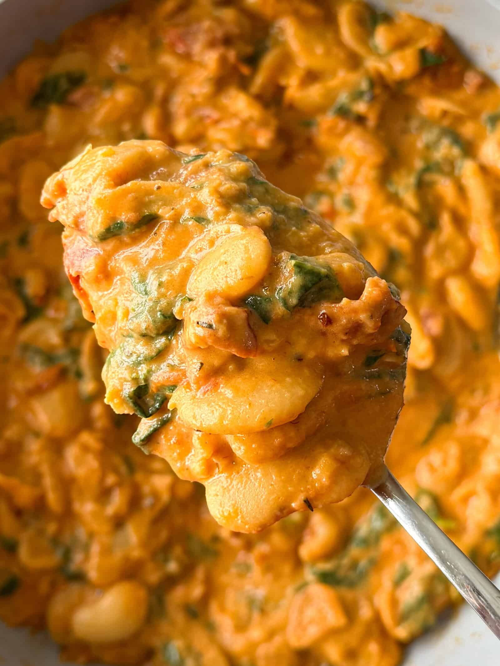 A spoonful of Marry Me Butter Beans in a creamy tomato sauce with spinach, held over a serving dish.