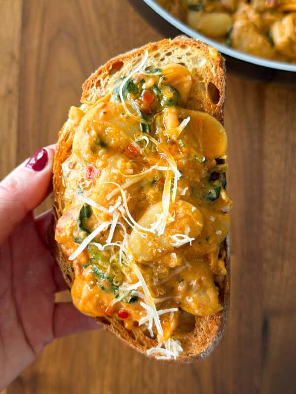 A hand holds a slice of toasted bread topped with creamy Marry Me butter beans, spinach, and shredded cheese.