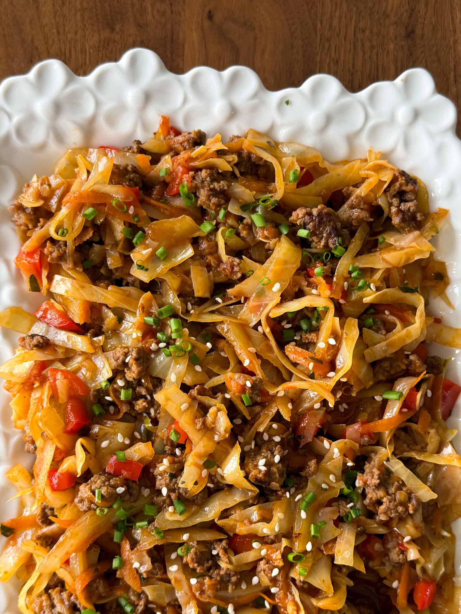 A white scalloped platter filled with a dirty cabbage stir-fry featuring ground meat, shredded cabbage, and red bell peppers, topped with sesame seeds and chopped chives.
