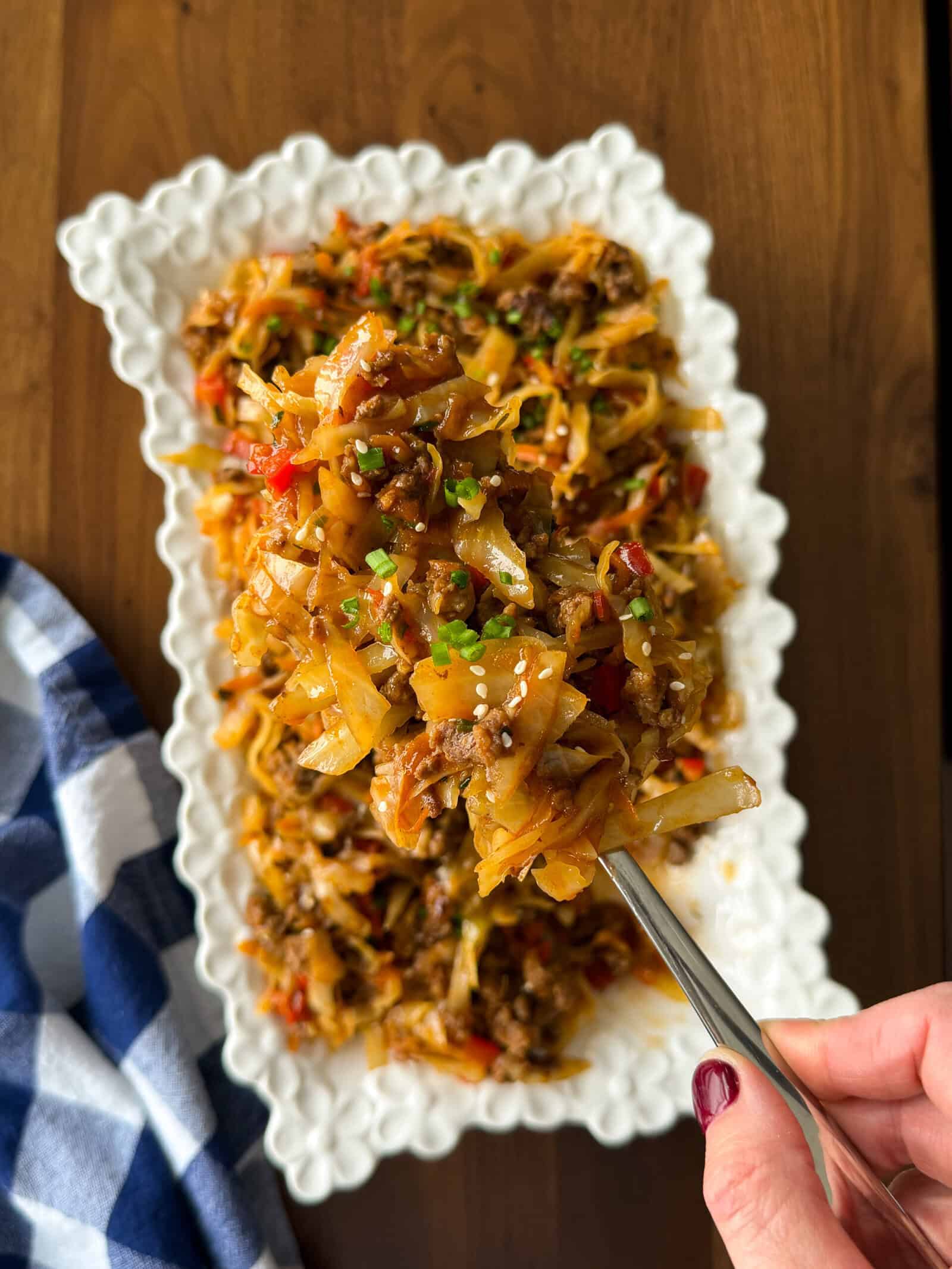 A serving spoon lifts a portion of dirty cabbage from a white decorative platter, revealing a mixture of sautéed cabbage, ground meat, and peppers.
