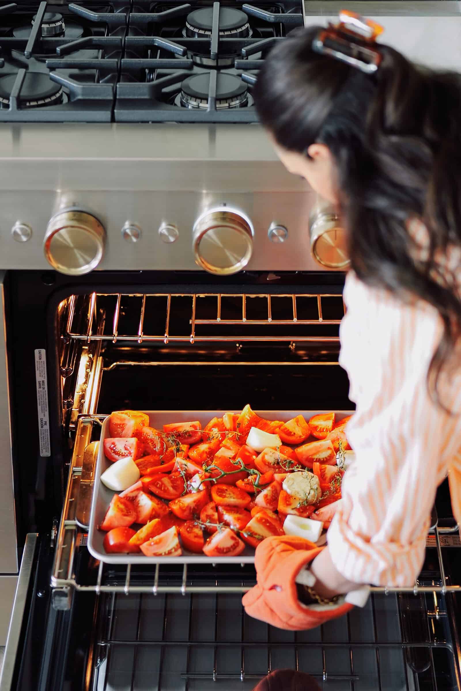 A person wearing an oven mitt slides a baking sheet of chopped tomatoes, onions, and herbs into an oven to prepare roasted tomato soup.