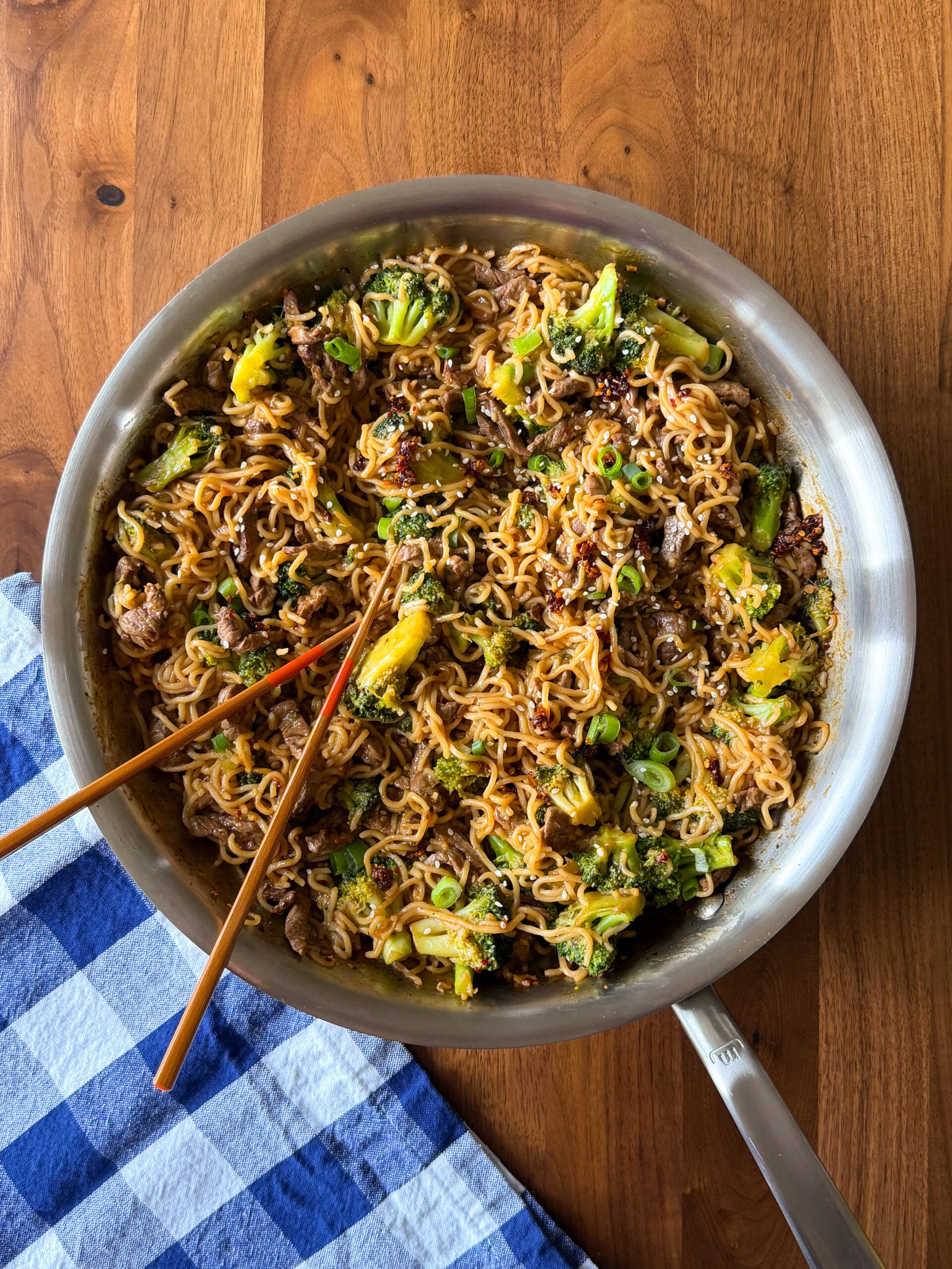 A stainless steel pan filled with beef and broccoli ramen stir-fry, with a pair of wooden chopsticks resting on top.