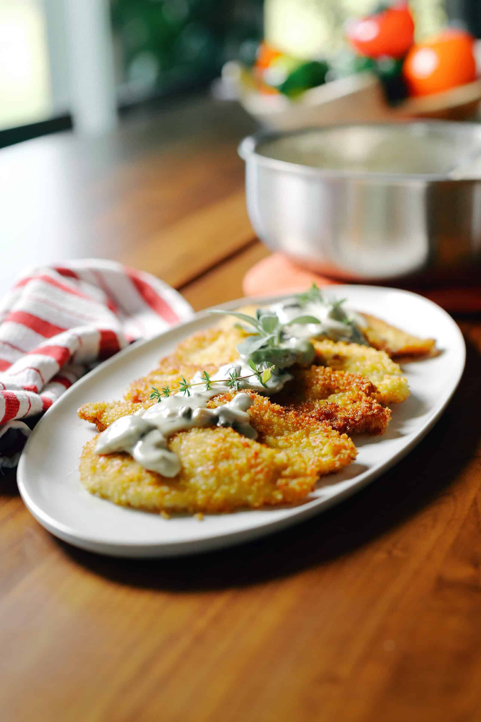 Breaded chicken cutlets topped with creamy sauce and herbs on an oval platter.
