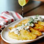A hand squeezes a lime wedge over a platter of breaded, pan-fried chicken cutlets topped with a creamy mushroom sauce.