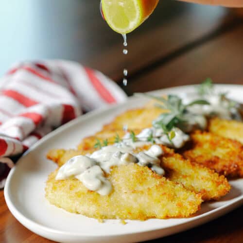 A hand squeezes a lime wedge over a platter of breaded, pan-fried chicken cutlets topped with a creamy mushroom sauce.