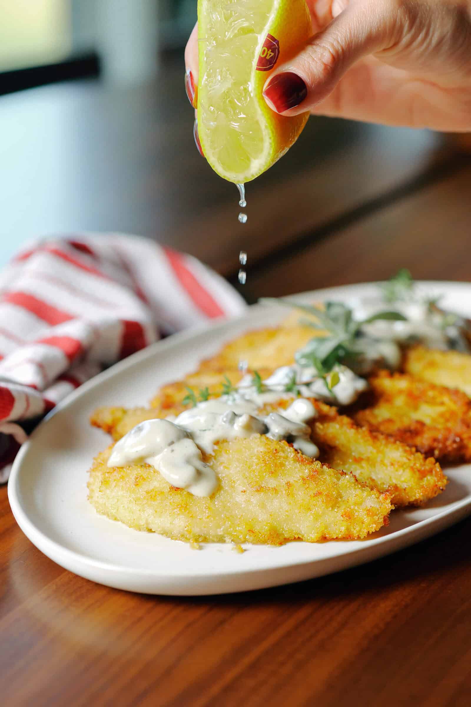 A hand squeezes a lime wedge over a platter of breaded, pan-fried chicken cutlets topped with a creamy mushroom sauce.
