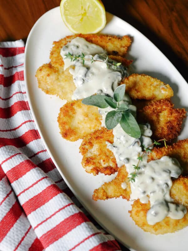 Breaded chicken cutlets topped with a creamy sauce and herbs on an oval platter with a lemon wedge.