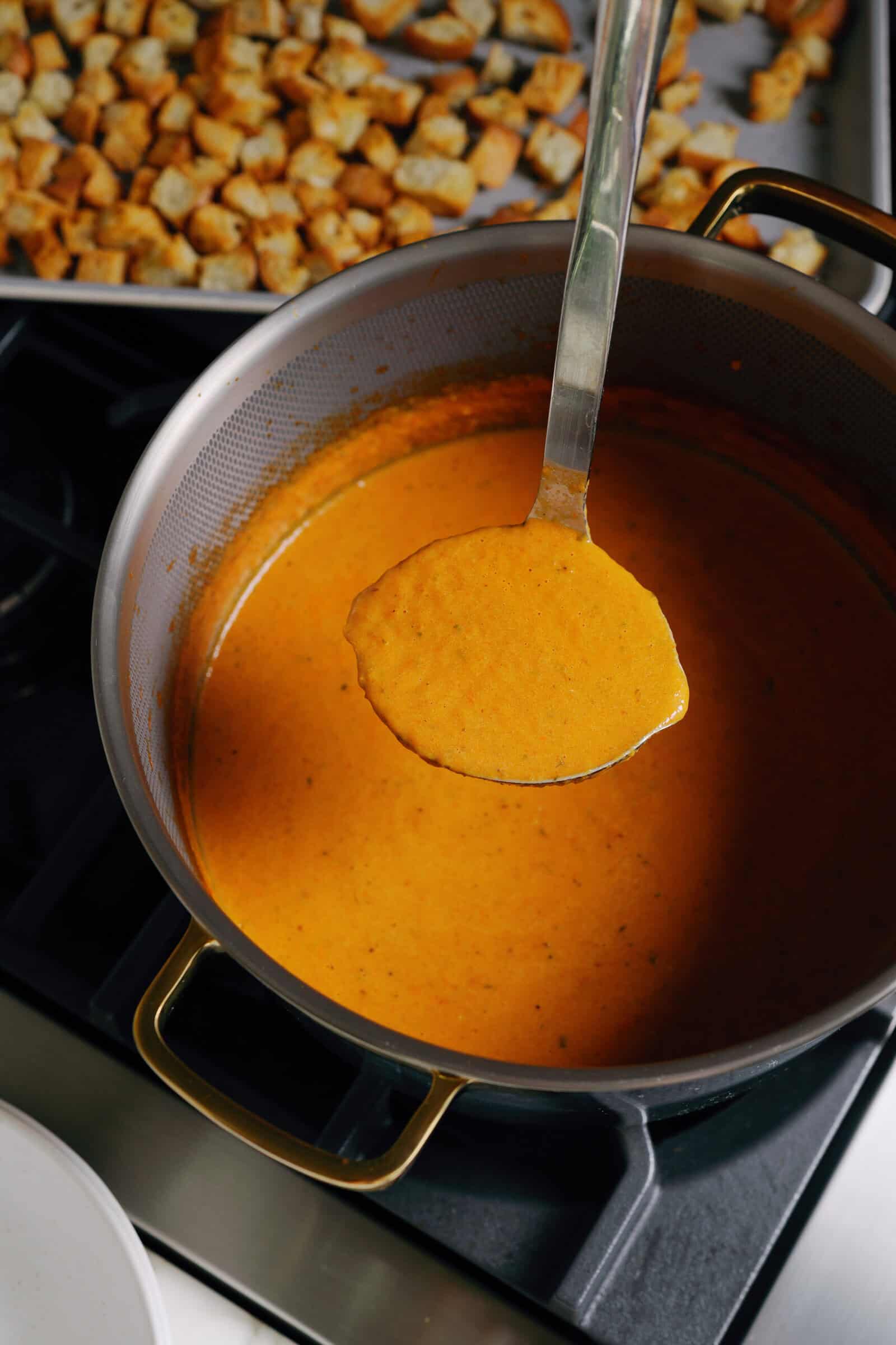 A metal ladle lifts orange roasted tomato soup from a pot on a stovetop, with a tray of croutons in the background.